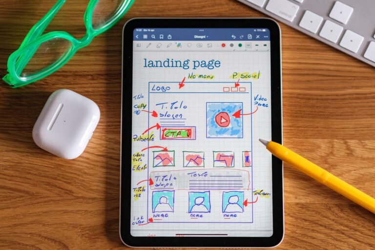 Tablet op een houten bureau met een handgetekende wireframe van een landingpagina voor een website. Rondom de tablet liggen een gele styluspen, draadloze oordopjes in een witte case, een groene bril en een toetsenbord. Op het scherm staan schetsen van navigatie, afbeeldingen, een video, tekstblokken en een call-to-actionknop, als voorbeeld van websiteontwerp en gebruikerservaring.