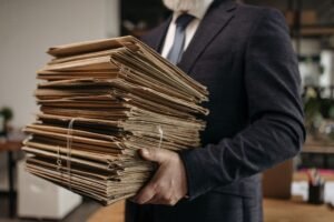 Persoon in een donker pak houdt een grote stapel papieren dossiers en documentmappen vast in een kantooromgeving. De documenten zijn gebundeld met elastieken en vertonen duidelijke gebruikssporen. Op de achtergrond zijn kantoorinrichting en werkplekken onscherp zichtbaar, wat de nadruk legt op de omvangrijke hoeveelheid papieren administratie.