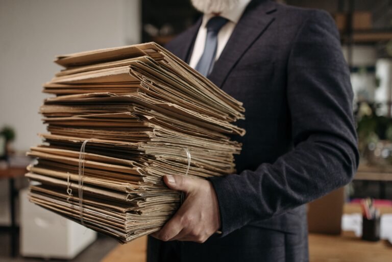 Persoon in een donker pak houdt een grote stapel papieren dossiers en documentmappen vast in een kantooromgeving. De documenten zijn gebundeld met elastieken en vertonen duidelijke gebruikssporen. Op de achtergrond zijn kantoorinrichting en werkplekken onscherp zichtbaar, wat de nadruk legt op de omvangrijke hoeveelheid papieren administratie.