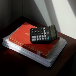 Rekenmachine ligt boven op een rood administratief boek dat op een stapel documenten op een donker houten bureau ligt. Zonlicht valt door een raam naar binnen en werpt sterke schaduwen op de witte muur en het bureau. De afbeelding roept een sfeer op van financiële administratie, boekhouding en kantoorwerk.