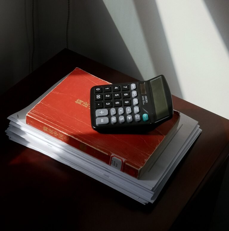 Rekenmachine ligt boven op een rood administratief boek dat op een stapel documenten op een donker houten bureau ligt. Zonlicht valt door een raam naar binnen en werpt sterke schaduwen op de witte muur en het bureau. De afbeelding roept een sfeer op van financiële administratie, boekhouding en kantoorwerk.