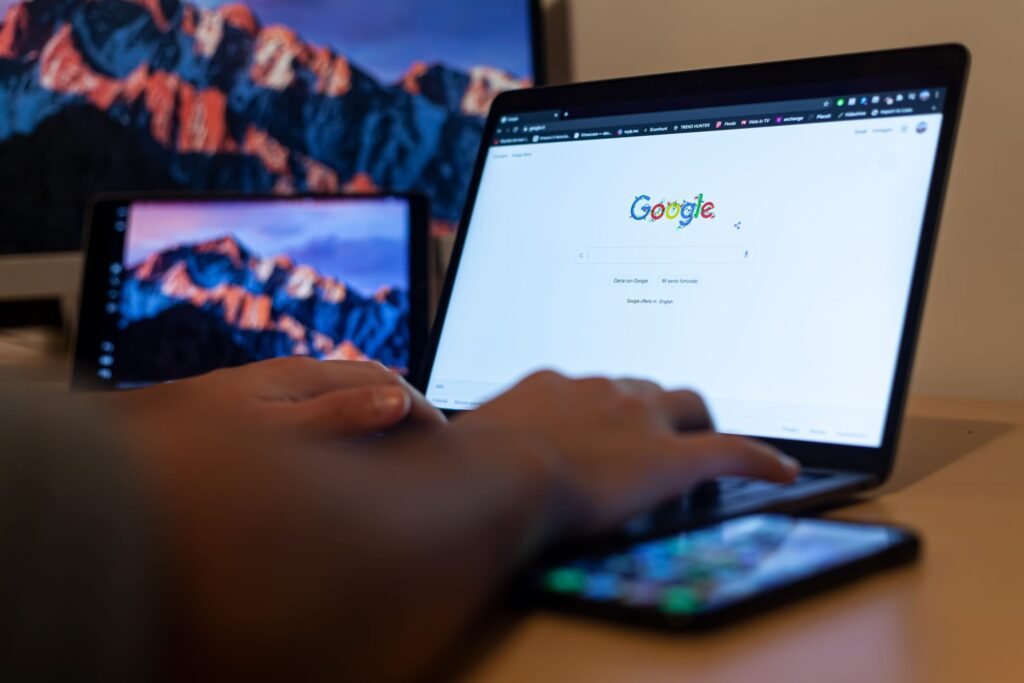 Persoon werkt op een laptop met de Google-zoekpagina open, naast een smartphone en meerdere schermen op een bureau.